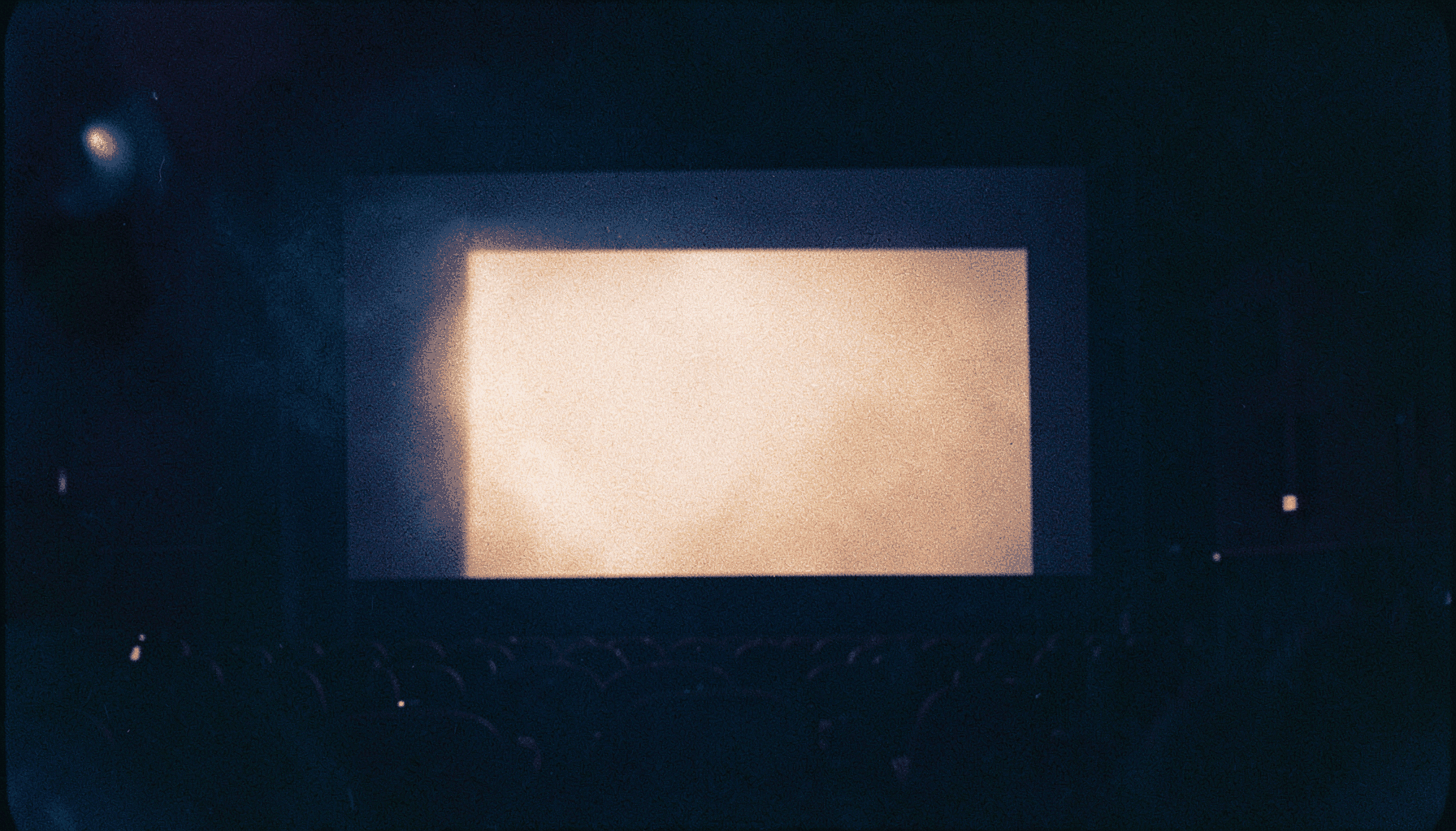 A warm, softly lit analog film photograph with visible grain showing a cinema screen glowing in a darkened theater, deep burgundy seats and amber wall sconces fading into shadow.