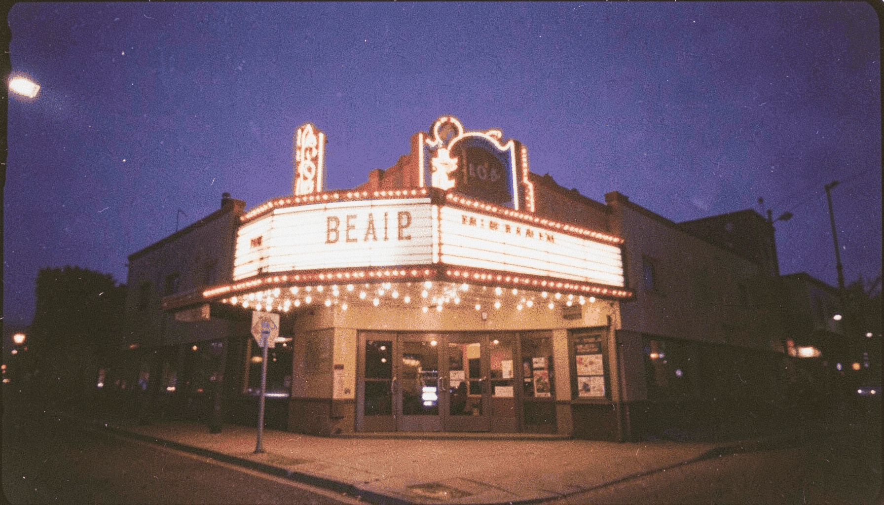 A grainy analog film photograph of a cinema marquee at dusk, warm amber bulbs glowing against a deep indigo sky, casting soft light onto the sidewalk below