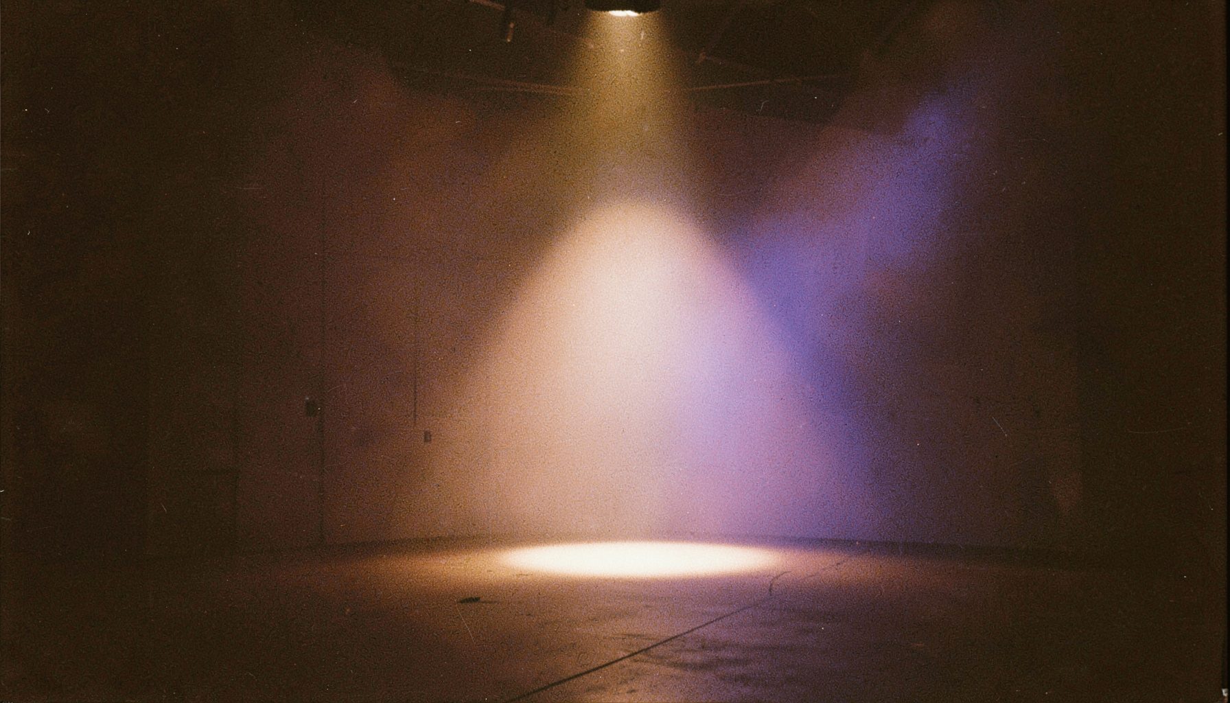 A grainy analog film photograph of a lone spotlight cutting through haze on an empty soundstage, warm amber tones against deep shadow