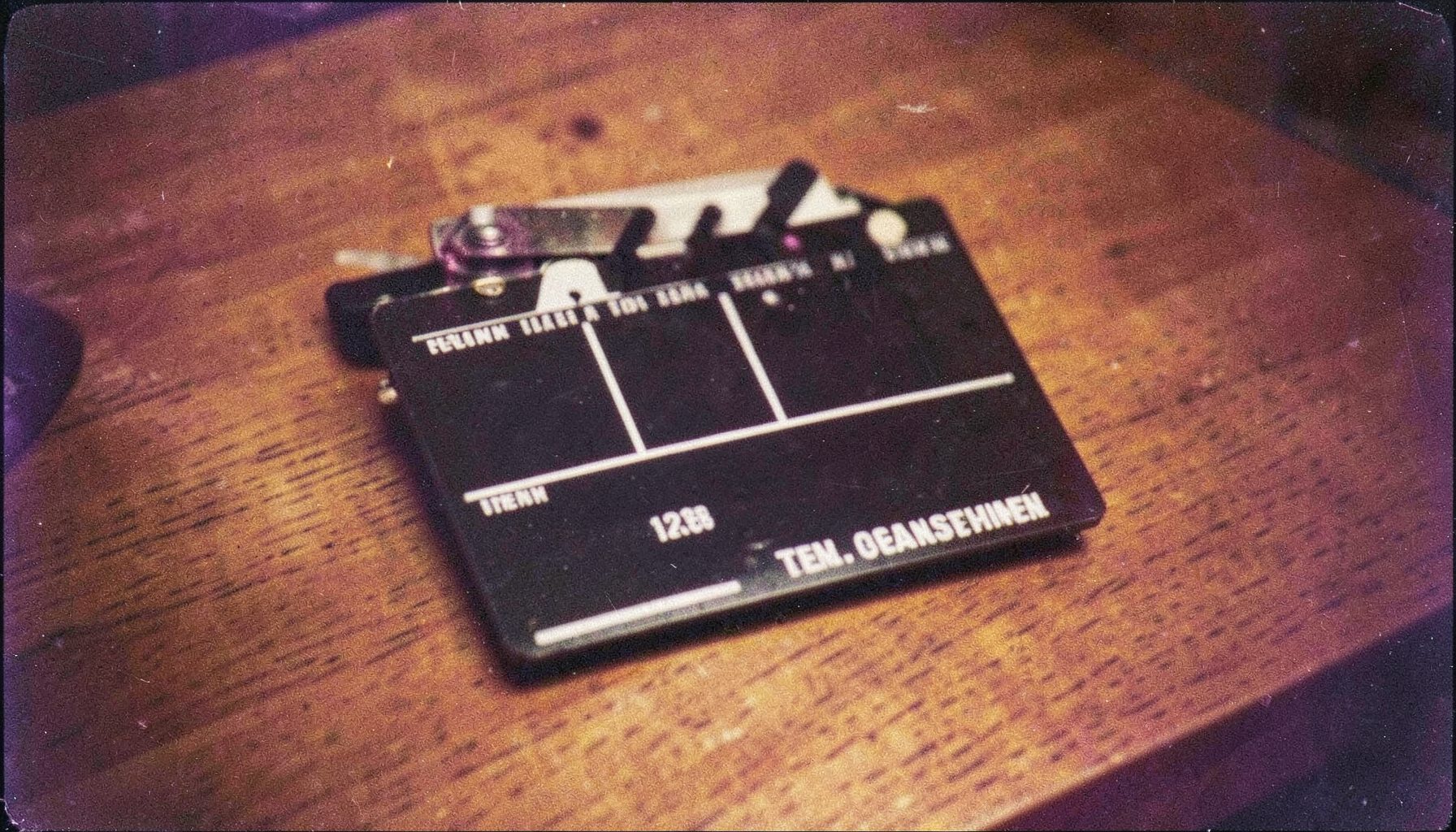 A grainy analog film photograph of a film slate resting on a wooden table under warm tungsten light, deep browns and muted golds