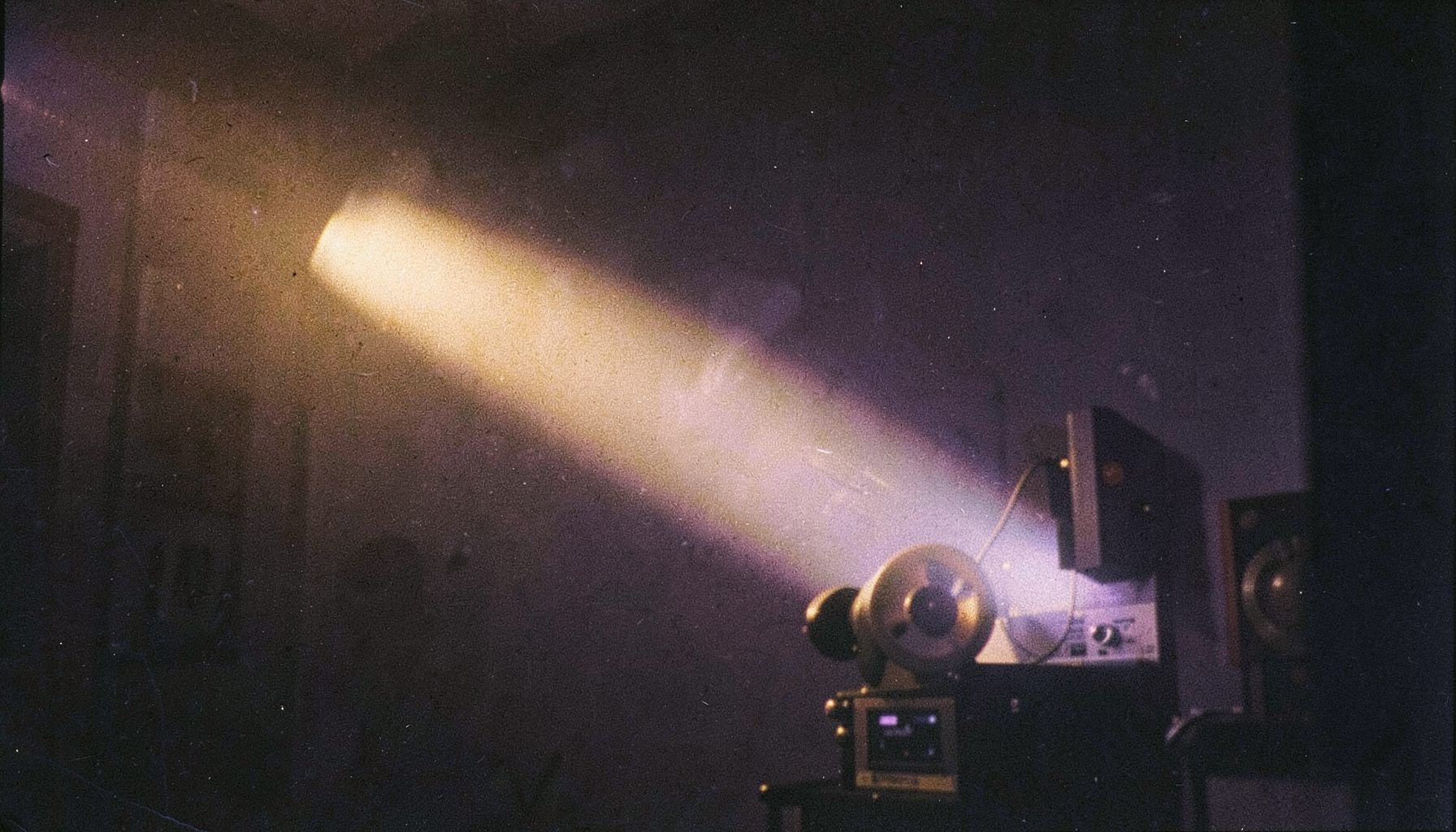 A grainy analog film photograph of a vintage film projector beam cutting through a darkened room, warm golden light with deep charcoal shadows