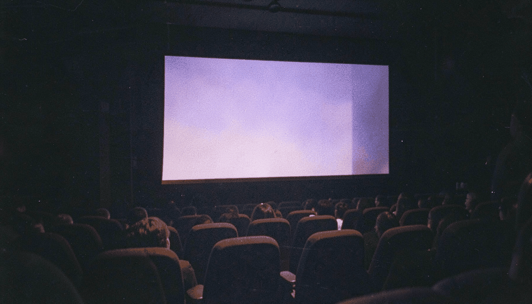 A grainy analog film photograph of a cinema screen glowing in a darkened theater, deep navy and warm amber light spilling across velvet seats, visible film grain